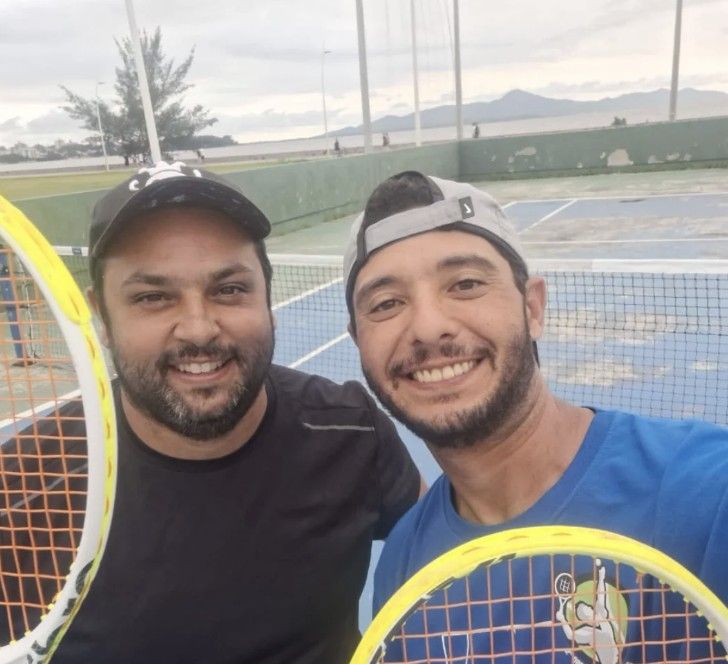 Rafael e um aluno sorrindo com as raquetes numa quadra à beira-mar, com a baía de Florianópolis ao fundo